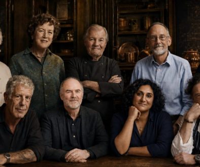 Editorial group portrait inspired by Auguste Escoffier, Julia Child, Jacques Pépin, Harold McGee, J. Kenji López-Alt, Samin Nosrat, Anthony Bourdain, Bill Buford, and Marco Pierre White, gathered around a rustic table in a warm, dark kitchen-library setting.